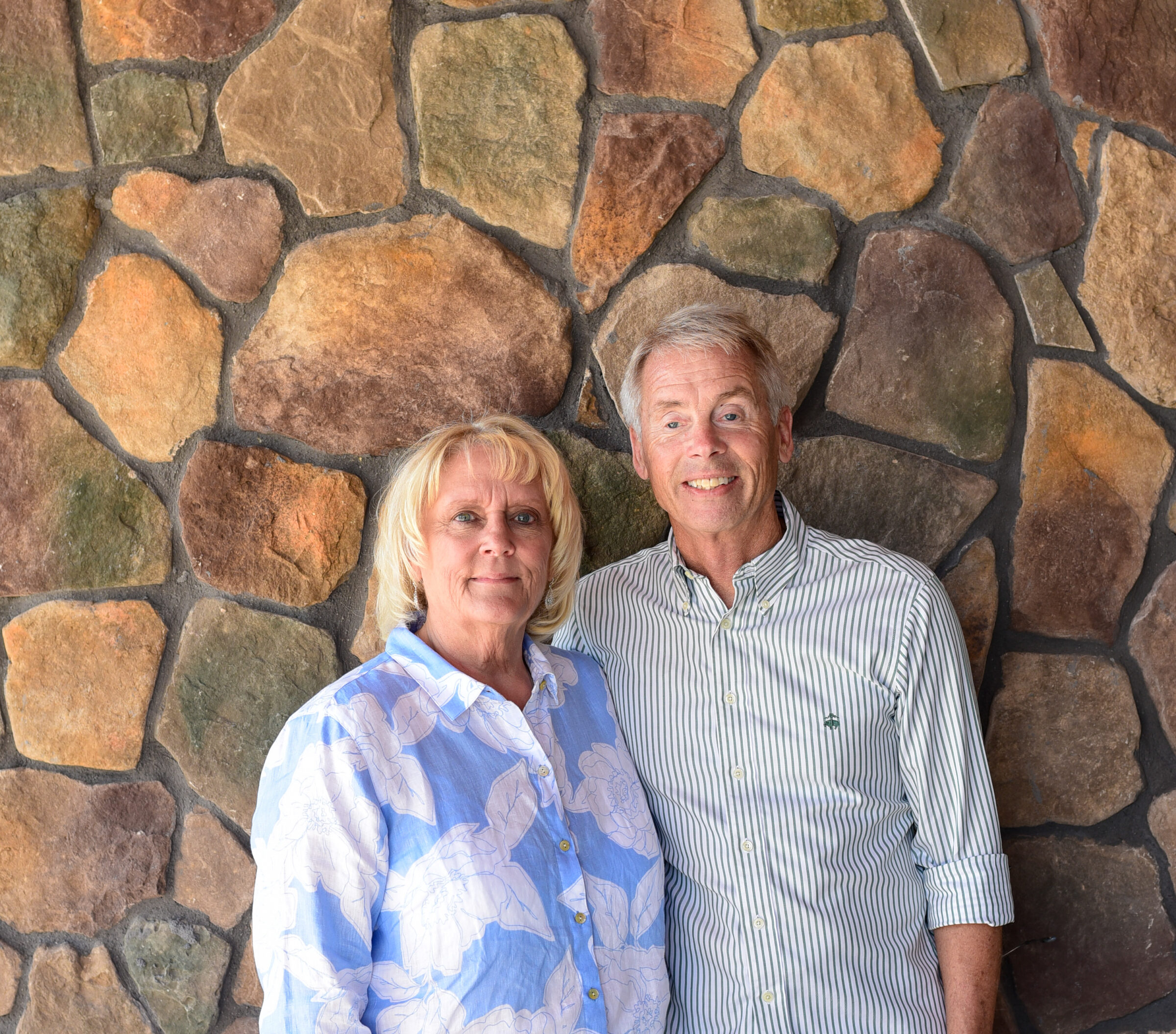 Frances and Tim Becker stand next to each other beside a stone wall.